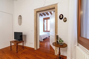 Un corridoio con pavimento in legno, una televisione su un tavolo, una porta che dà su una camera da letto e una finestra con tende.