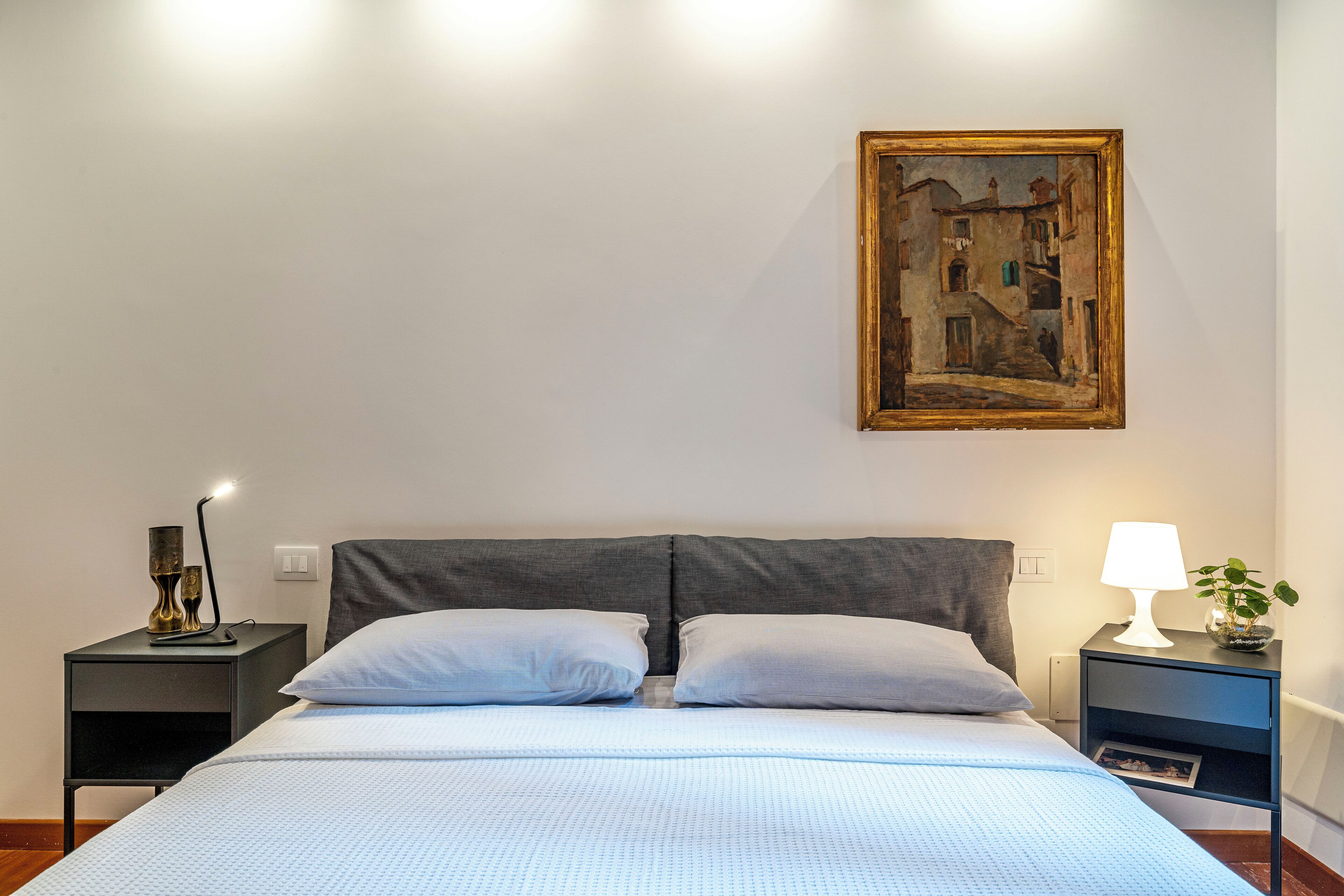 Uma cama bem arrumada com cabeceira cinza, duas mesas de cabeceira com abajures, um quadro na parede e uma pequena planta sobre uma das mesas.
