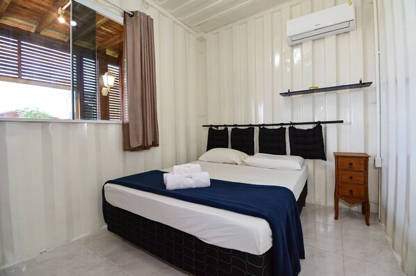 Una cama bien hecha con una colcha azul y blanca, una mesita de noche de madera con cajones, un estante en la pared y un equipo de aire acondicionado.