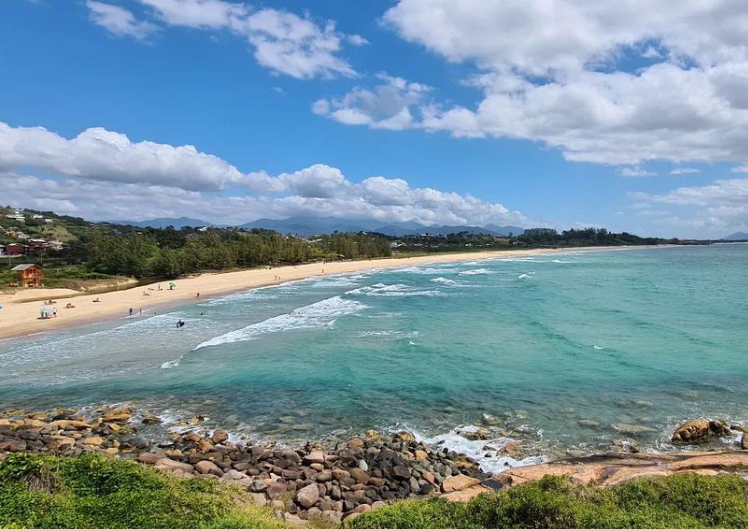 A coastal beach scene with clear turquoise waters, a sandy shore, and a rocky foreground.