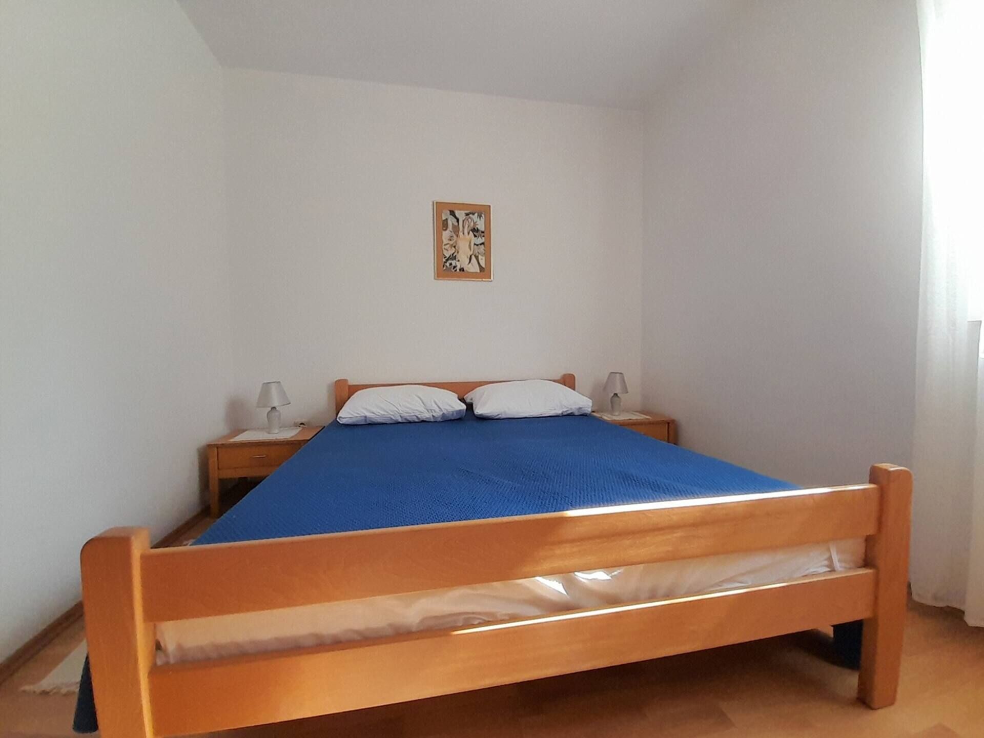 A single-bed room with a wooden frame, a blue mattress, white pillows, two bedside tables with lamps, and a framed picture on the wall.