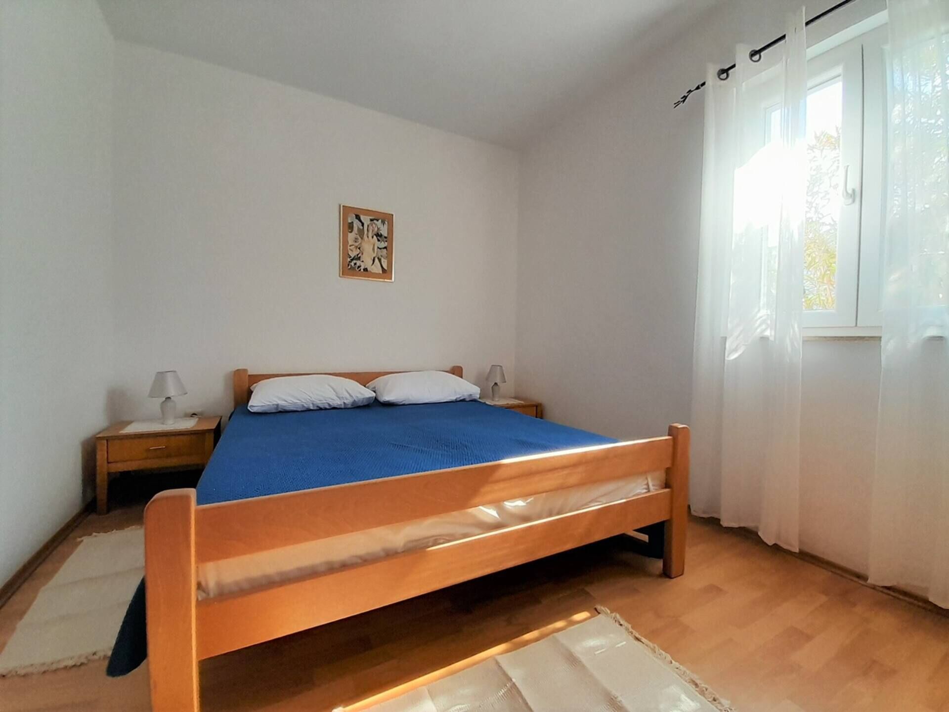 Een slaapkamer met een houten bed, een nachtkastje, een lamp, een raam met gordijnen en een schilderij aan de muur.