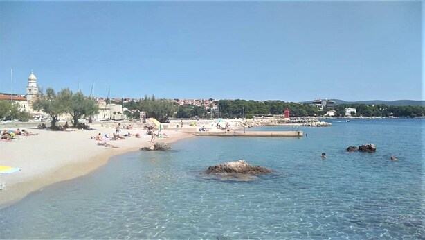 A beach with clear water, a sandy shore, and a few people. There is a lighthouse in the background and a building with a tower.
