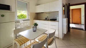 A compact kitchen with white cabinets, a small dining table with cushioned chairs, a window with blinds, and a door leading to another room.