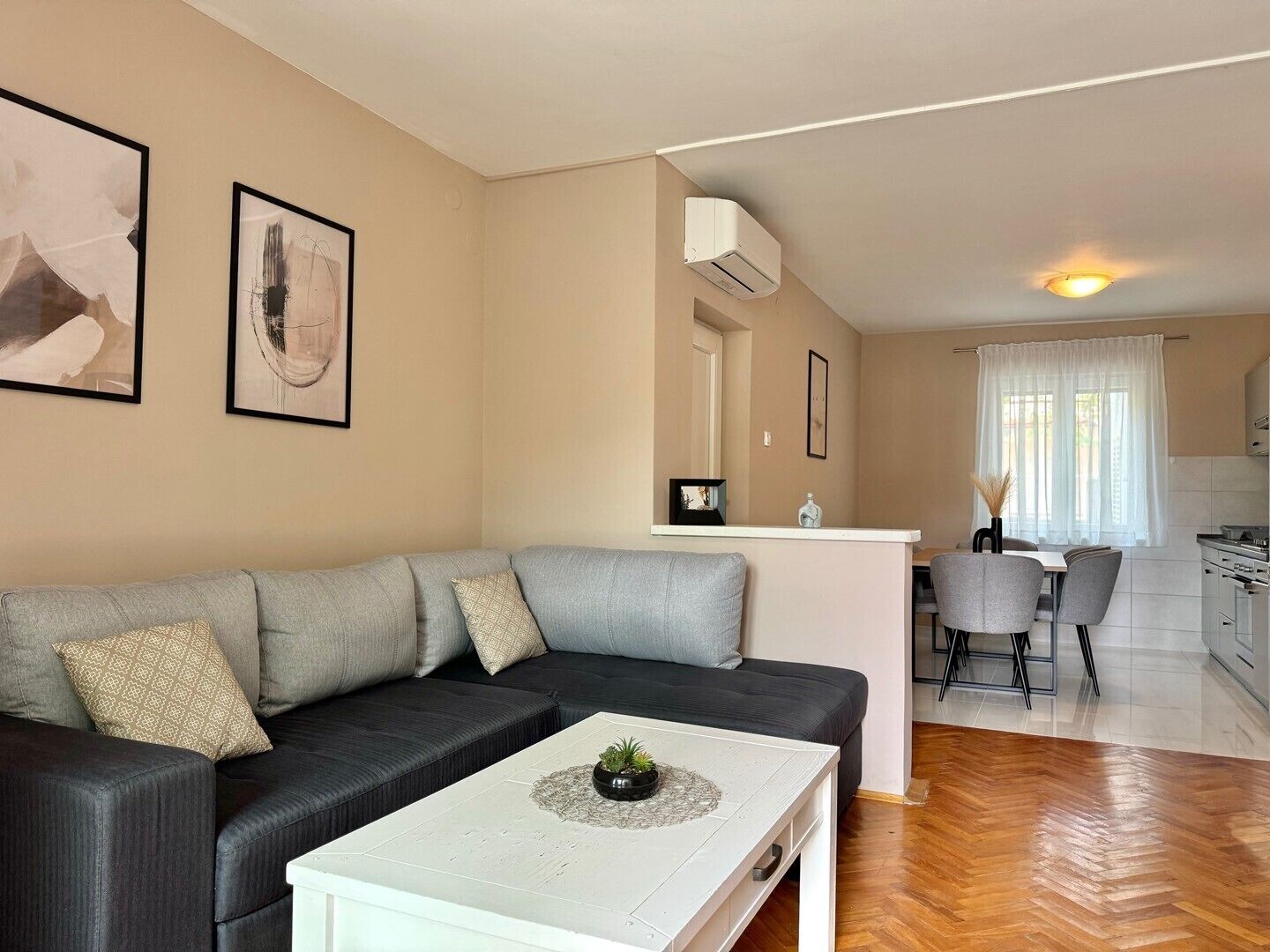 A modern living room with a grey sofa, a white coffee table with a plant, a kitchen area with a dining table and chairs, and artwork on the walls.
