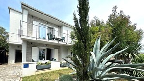 A modern two-story building with a balcony, outdoor seating, and a garden with large agave plants. - Apartment DiSente °1 in Krk Family-friendly, Close to the center, Doga allowed (Krk)