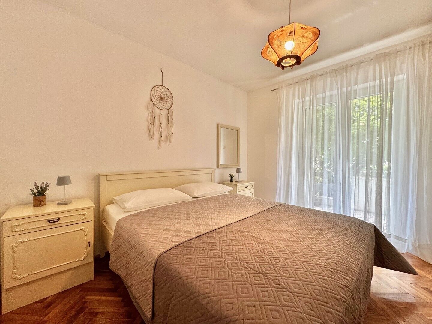 Une chambre à coucher avec un lit, des tables de chevet, une commode, un attrape-rêves, un luminaire suspendu et des rideaux légers.