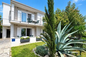 Un bâtiment à deux étages doté d’un balcon, d’un espace extérieur aménagé pour s’asseoir et d’un jardin bien entretenu où poussent différentes espèces de plantes.