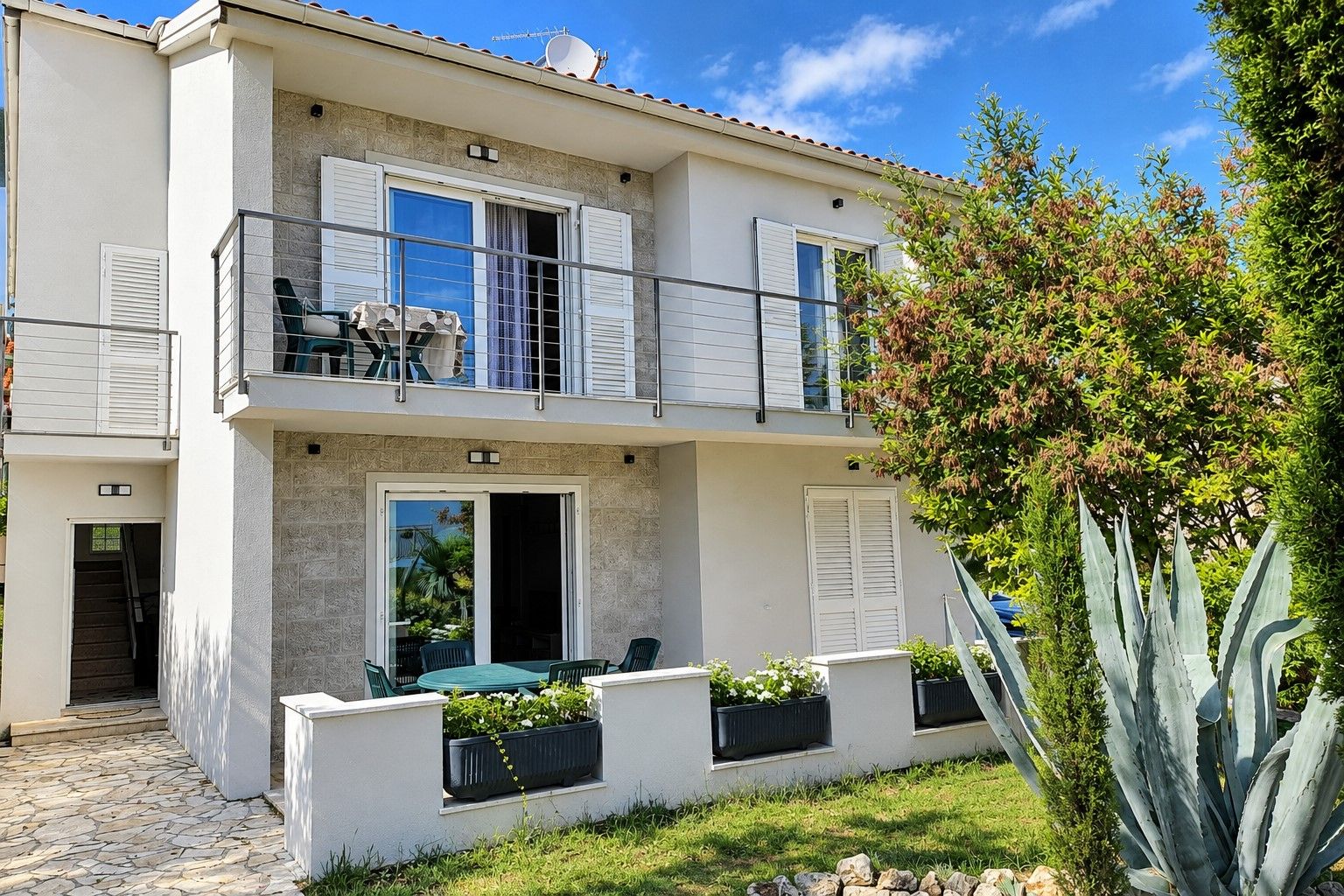 A two-story building with a balcony, a dining area with outdoor furniture, and a garden with a large agave plant.