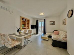 Uma sala de estar com sofá, mesa de jantar com cadeiras, estante de livros, televisão e janela com cortinas.