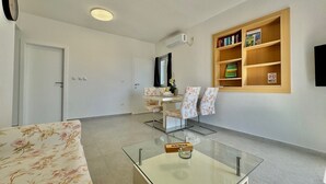 A hotel lobby with a sofa, a glass coffee table, a small table with a box, a bookshelf, and a door.