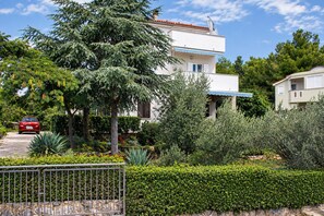 A two-story building with a balcony, surrounded by greenery and a parked car.