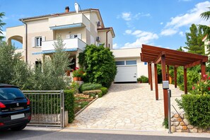A two-story residential building with a stone-paved entrance, a covered wooden pergola, and a car parked in front.