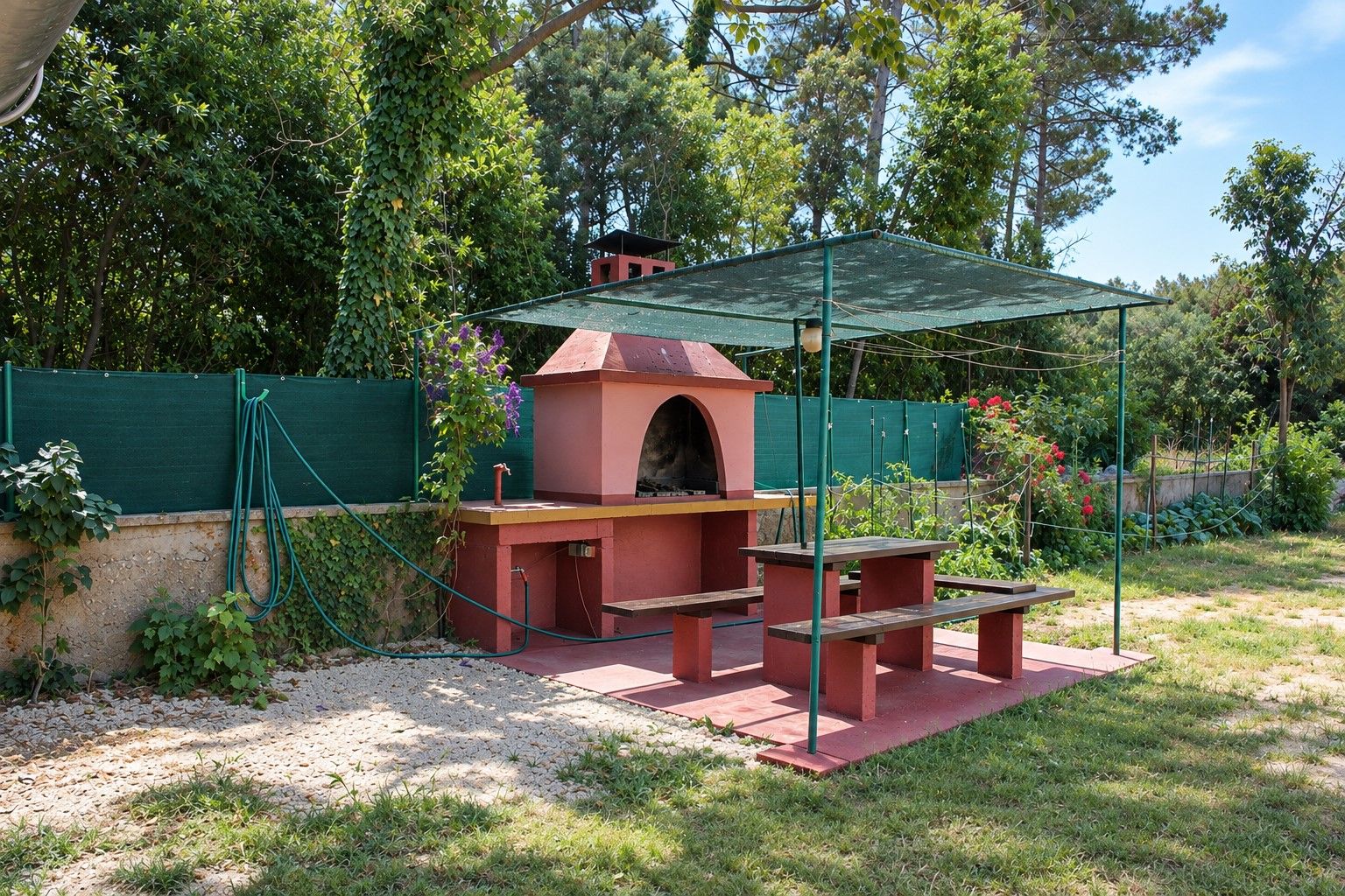 An outdoor barbecue area with a brick oven, a covered seating area, and a green fence.