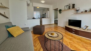 A modern living room with a sofa, ottoman, round coffee table, TV, and a kitchen in the background. - Apartment DiSente °2 in Krk, family-friendly, close to the center, pets allowed, sea view (Krk)