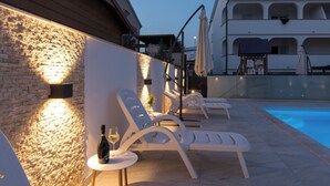 A poolside area with lounge chairs and a bottle of wine, a stone wall, and a building with arched windows in the background.