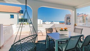 A balcony with a table and chairs, a hanging chair, and a view of the sea and buildings.