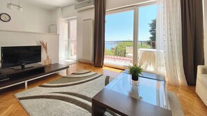 Un soggiorno moderno con una televisione a schermo piatto, un tavolino in vetro e vista sul mare attraverso ampie finestre.