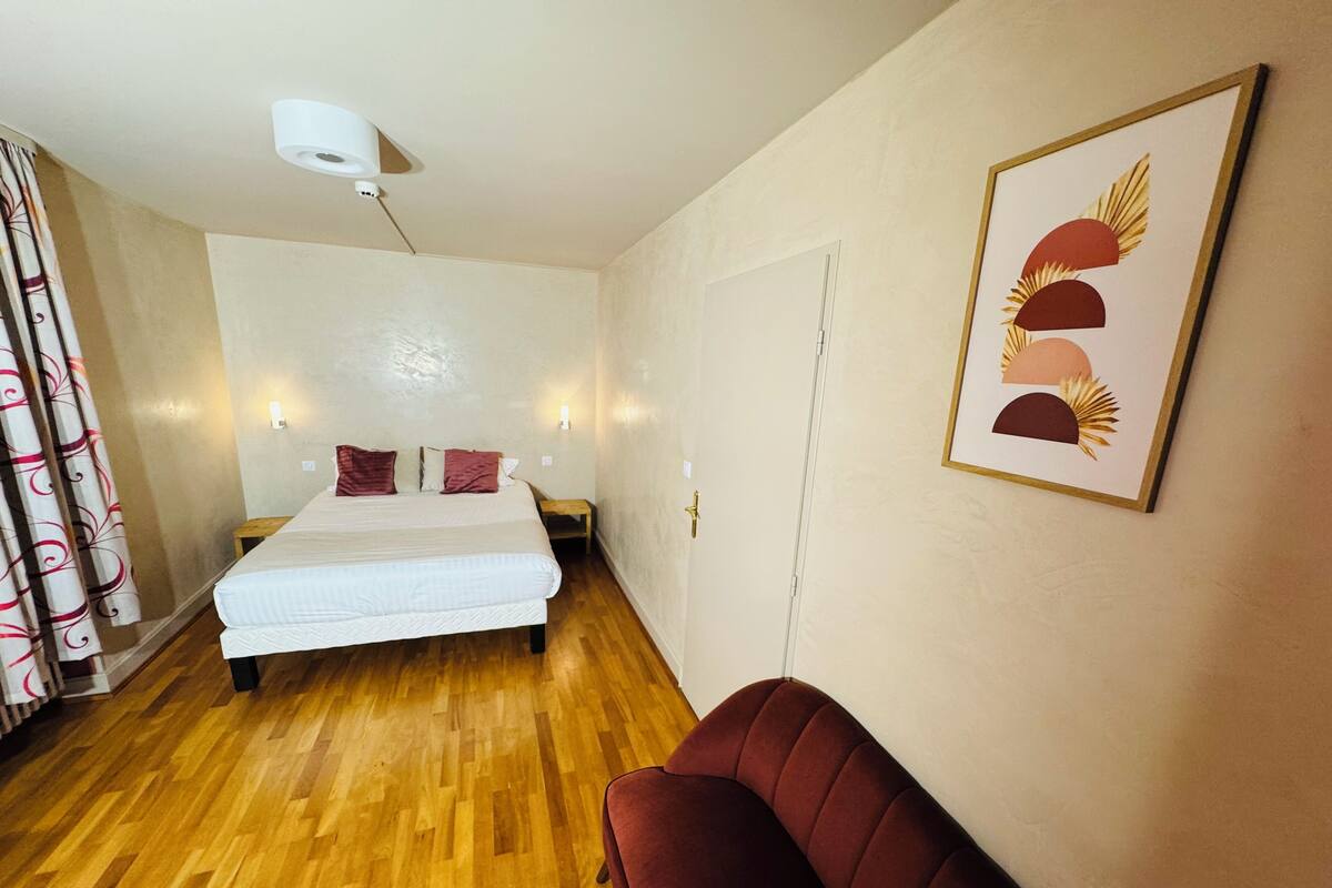 Ein Schlafzimmer mit einem Bett, Holzboden, einem roten Sessel, einer Wandleuchte und einem gerahmten Bild.