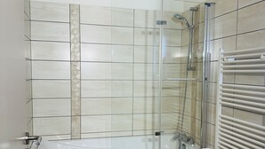 Une salle de bain moderne avec une douche à parois de verre, un tabouret en bois sur lequel sont posées des serviettes et un lavabo blanc.