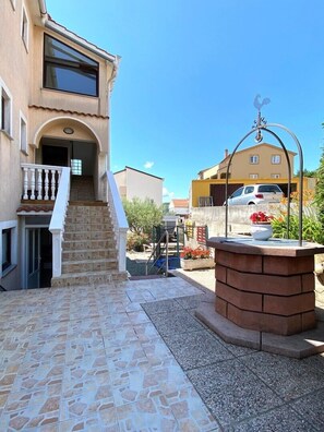 A residential building with a stone staircase leading to a door, a small garden area with a decorative fountain, and a parked car in the background. - Family-friendly apartment Lilly with garden barbecue and trampoline (Krk)