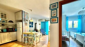A spacious kitchen with a dining area, a small table, and a chair. There is a refrigerator, a microwave, and a sink. The room has a tiled floor and a window with blue curtains.