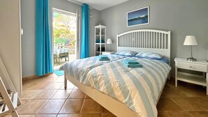 A bedroom with a bed, two bedside tables, a nightstand, a chandelier, a window with curtains, a door, and a view of the outdoors.