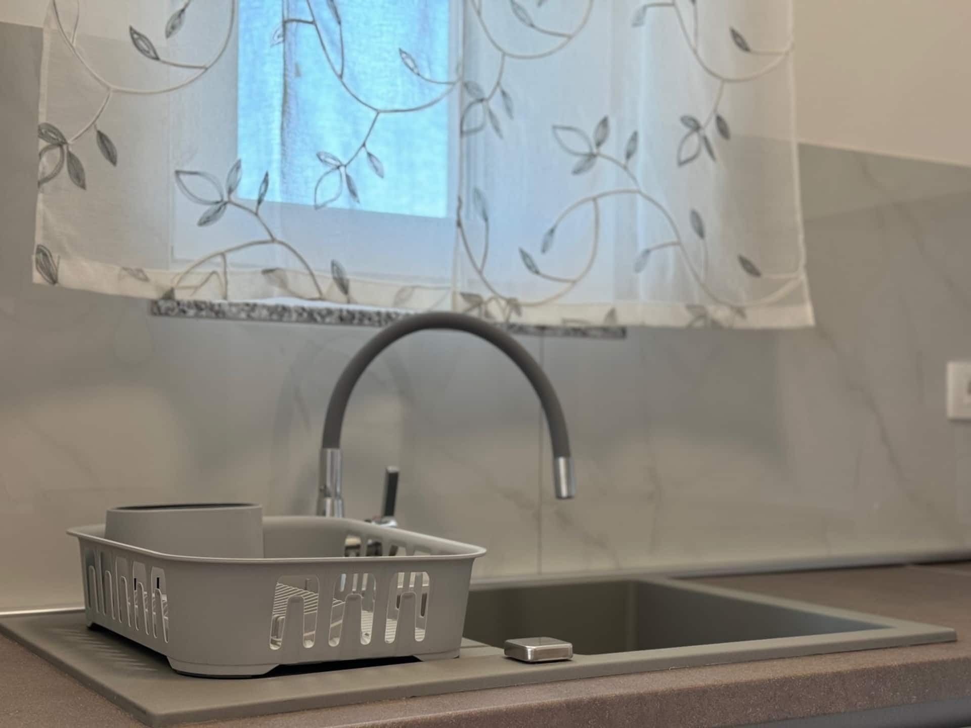 Une cuisine avec un évier moderne, un panier gris et une fenêtre ornée d’un rideau à motifs floraux.
