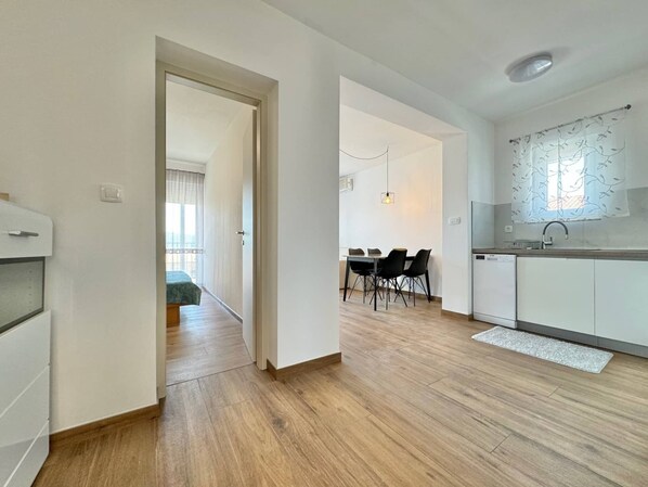 A modern kitchen with white cabinets, a dining area with a table and chairs, and a hallway leading to a bedroom.