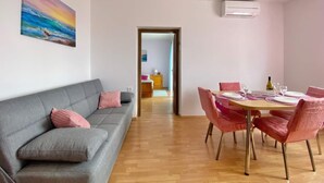 Un salon moderne avec un canapé gris, des coussins roses, une table à manger avec des chaises roses et un tableau représentant un coucher de soleil sur l’océan, accroché au mur.