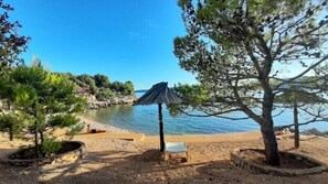 Une plage offrant une vue dégagée sur la mer, une zone sablonneuse avec un banc et un arbre sous lequel se trouve un parasol noir.