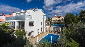 Un edificio moderno blanco con piscina, rodeado de vegetación y otros edificios.