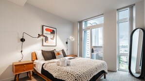 A modern bedroom with a large bed, bedside tables, a wall-mounted lamp, a framed abstract artwork, and a large window with a view of buildings. - 2BR | Gym, Lounge | Bellevue | Sophari Downtown E (Bellevue)