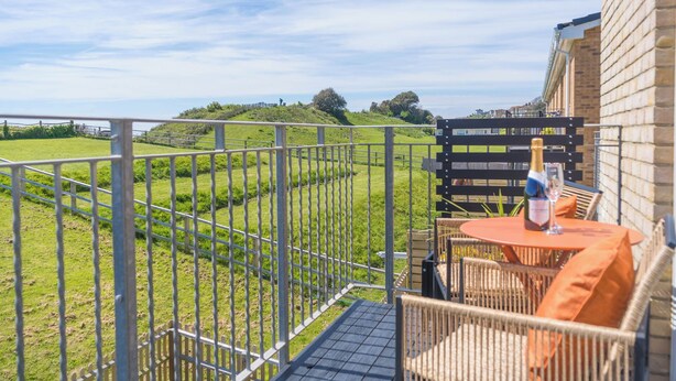 Ein Balkon mit Blick auf ein grünes Feld, ein Tisch mit Flasche und Glas sowie ein Backsteinhaus.