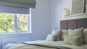 Ein Schlafzimmer mit großem Fenster, einem Bett mit gemusterten Kissen und einem grauen Kopfteil sowie einem Heizkörper.