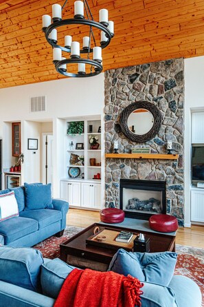 A living room with a stone fireplace, blue sofas, a coffee table, and a chandelier. - Red White & WTG: Hot Tub, 5 Bedrooms! (Wintergreen Resort)