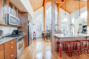 A spacious kitchen with wooden cabinets, a large island with stools, and a view of the outdoors through large windows. - Red White & WTG: Hot Tub, 5 Bedrooms! (Wintergreen Resort)