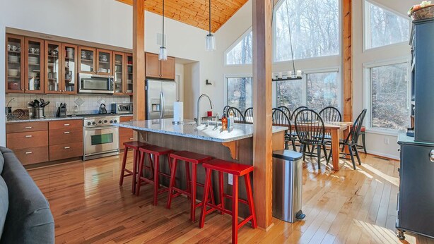 A spacious kitchen with wooden beams, a central island with stools, and large windows offering a view of the outdoors. - Red White & WTG: Hot Tub, 5 Bedrooms! (Wintergreen Resort)