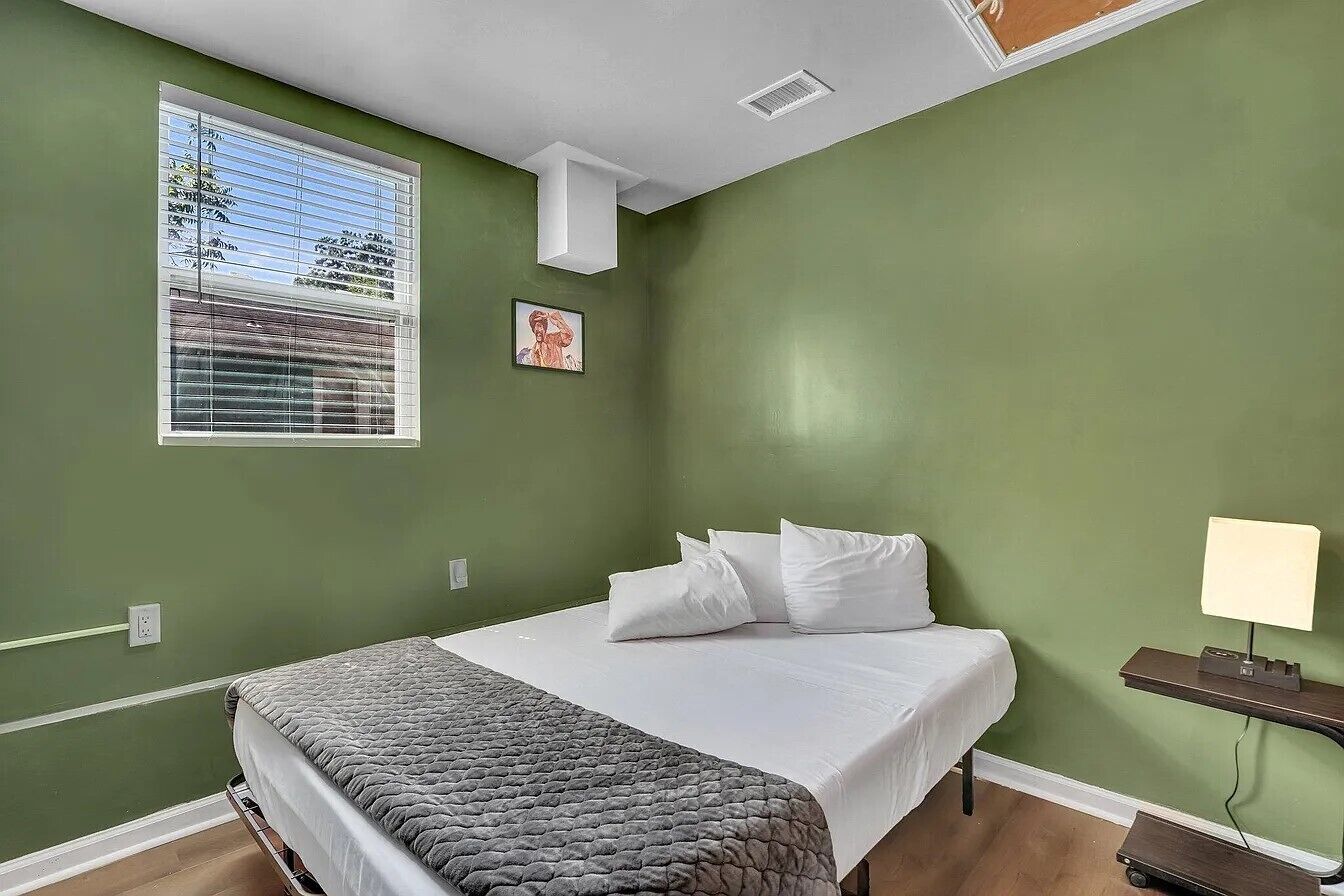 Ein Schlafzimmer mit einem Bett, einem Fenster mit Jalousien, einer Wandleuchte und einem kleinen Bild.