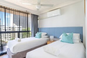 Ein Hotelzimmer mit zwei Betten, einem Deckenventilator, einer Klimaanlage und Blick auf Gebäude durch das Fenster.