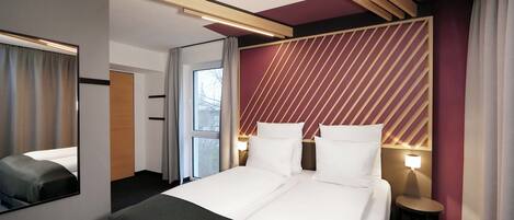 A hotel room with a bed, a mirror, a bedside table with a lamp, and a view of the outside through a window.