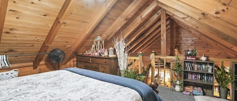 Un dormitorio acogedor en el ático, con techo inclinado, cama con colcha estampada en blanco y gris, cómoda de madera, estantería y un ventilador.