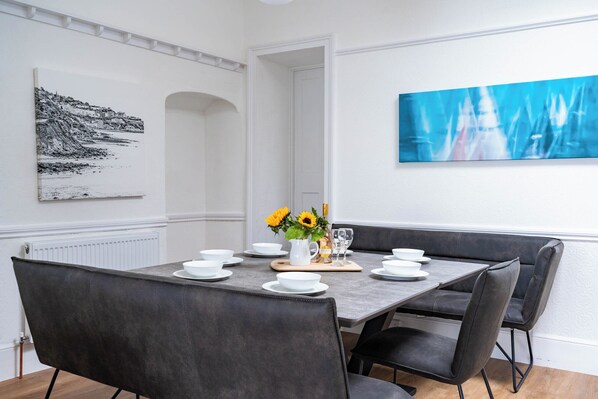 A dining area with a rectangular table set for four, surrounded by dark chairs. There is a white radiator below the table, and a large abstract painting is visible on the wall to the right. - Breathtaking 180° Ocean Views (Ventnor)