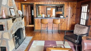 Interior acogedor de una cabaña con chimenea de piedra, paredes de madera, una zona de bar con taburetes y un sillón de cuero.