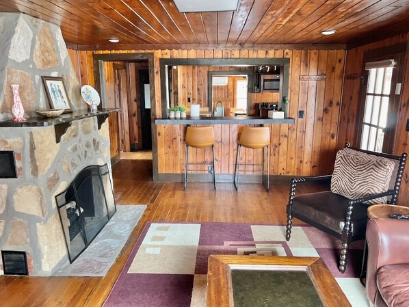 A cozy cabin interior with a stone fireplace, wooden walls, a bar area with stools, and a leather armchair.