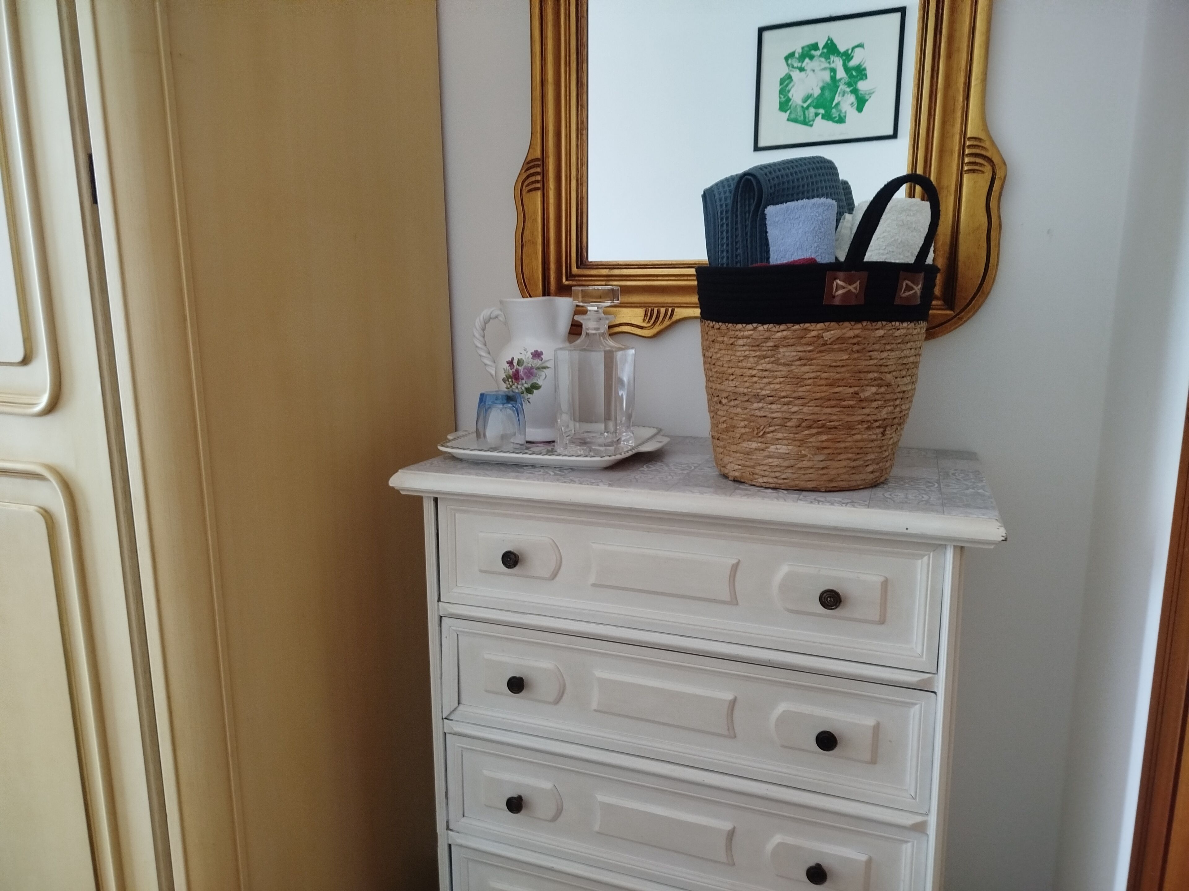A white dresser with drawers, a mirror with a gold frame, and a wicker basket with towels.