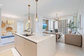 A modern kitchen with a central island, a dining area with a white table and chairs, and a living area with a sofa and a TV.