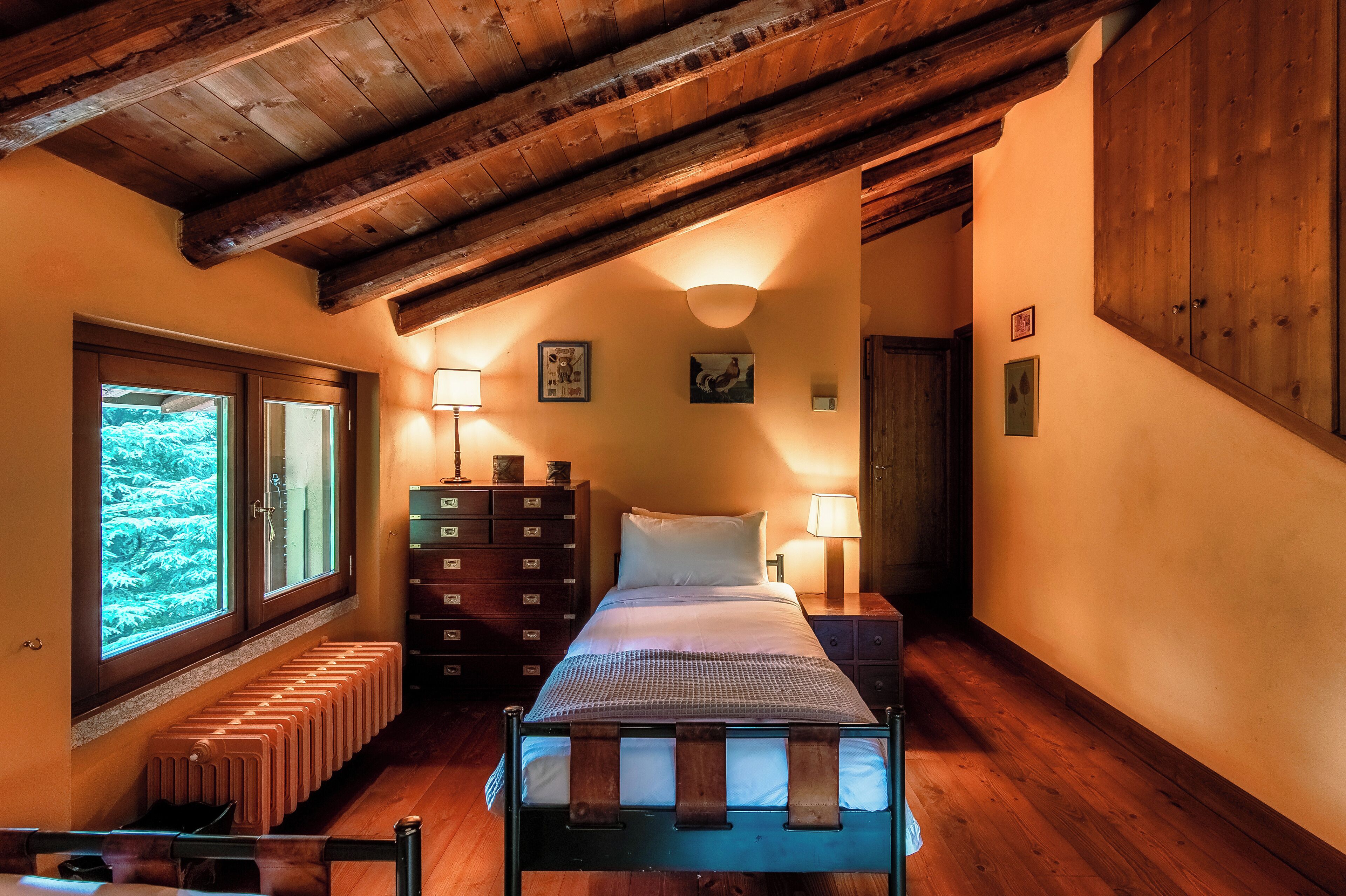 A bedroom with a wooden ceiling, a bed with a headboard, a nightstand, a dresser, and a window with a view of greenery.
