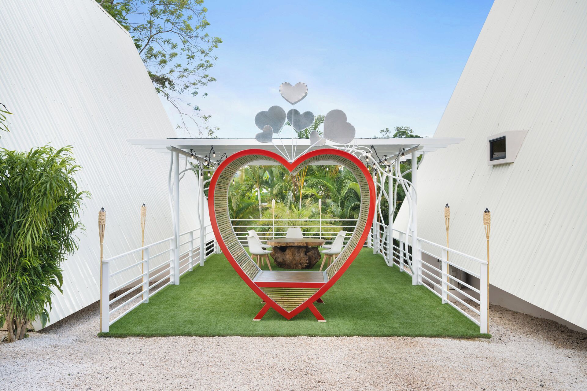 A heart-shaped structure with a table and chairs in a white-walled area with artificial grass.