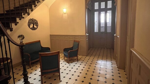 Un corridoio con pavimento a scacchi bianco e nero, una scala in legno, un divano blu e due sedie di legno.